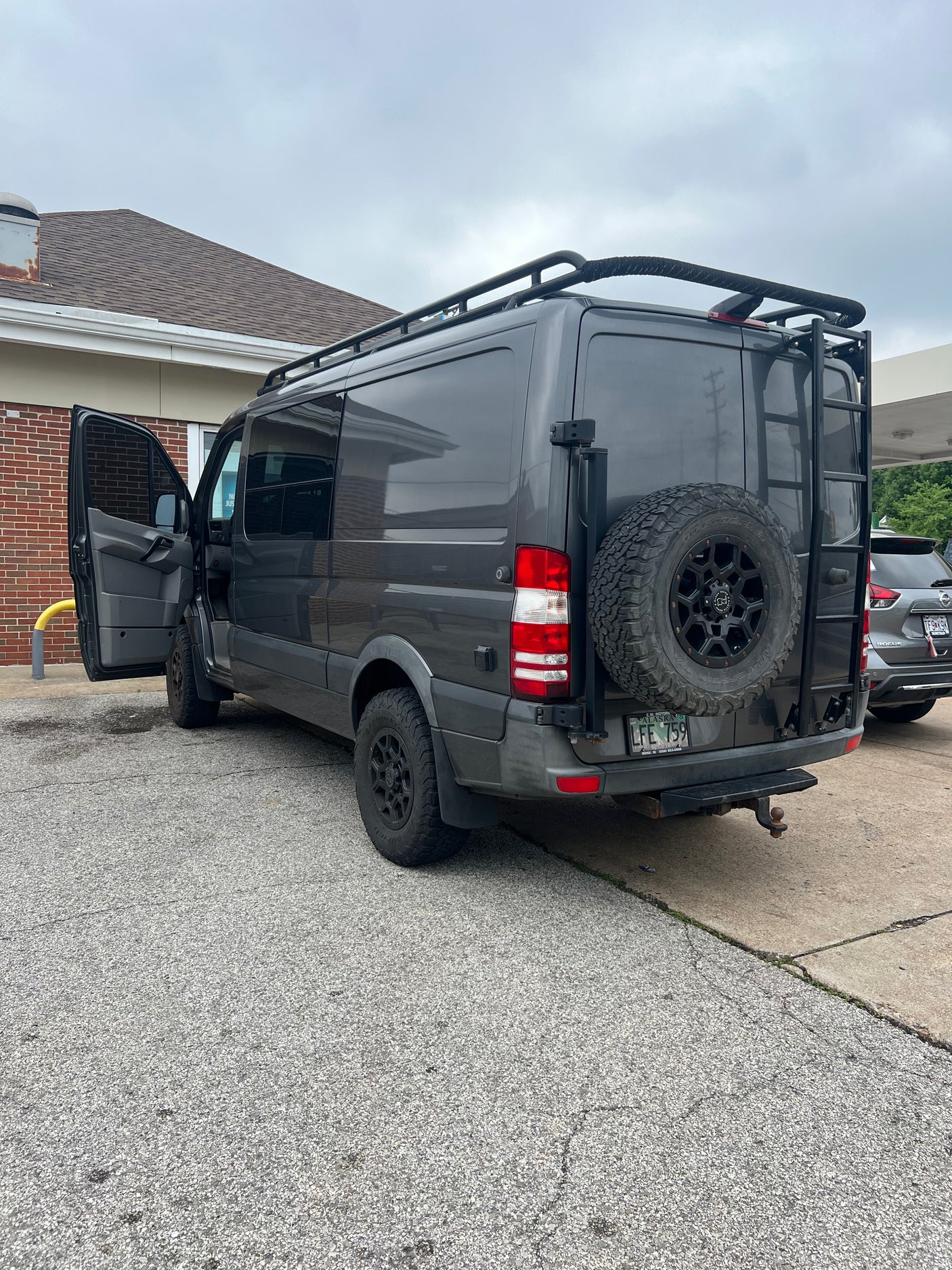 Grey Sprinter RV rental, equipped for adventure with roof rack, ladder, and rear spare tire, parked on asphalt.