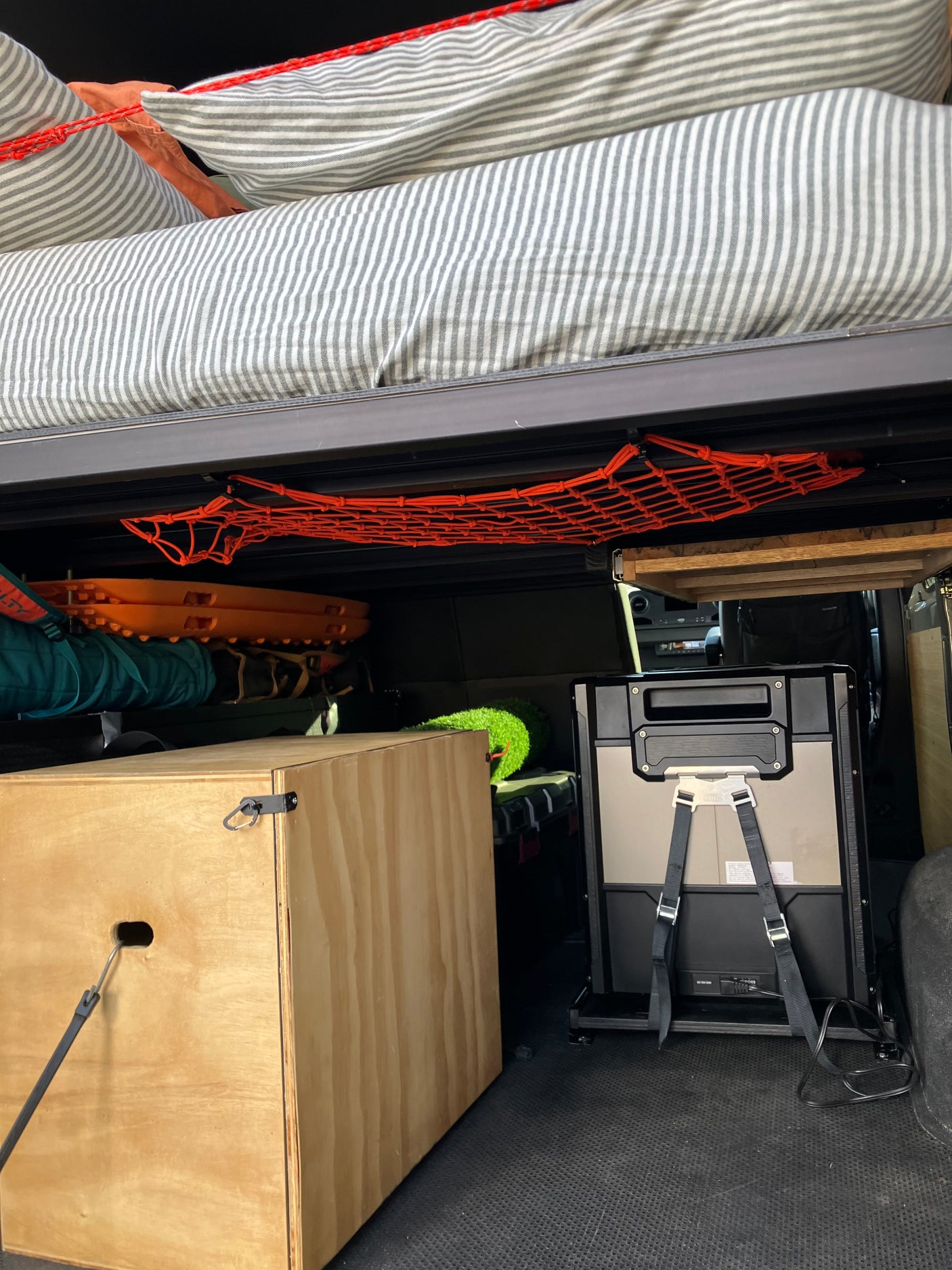 RV rental interior: Under-bed storage with striped bed, orange gear net, traction boards, wooden box, and portable fridge.