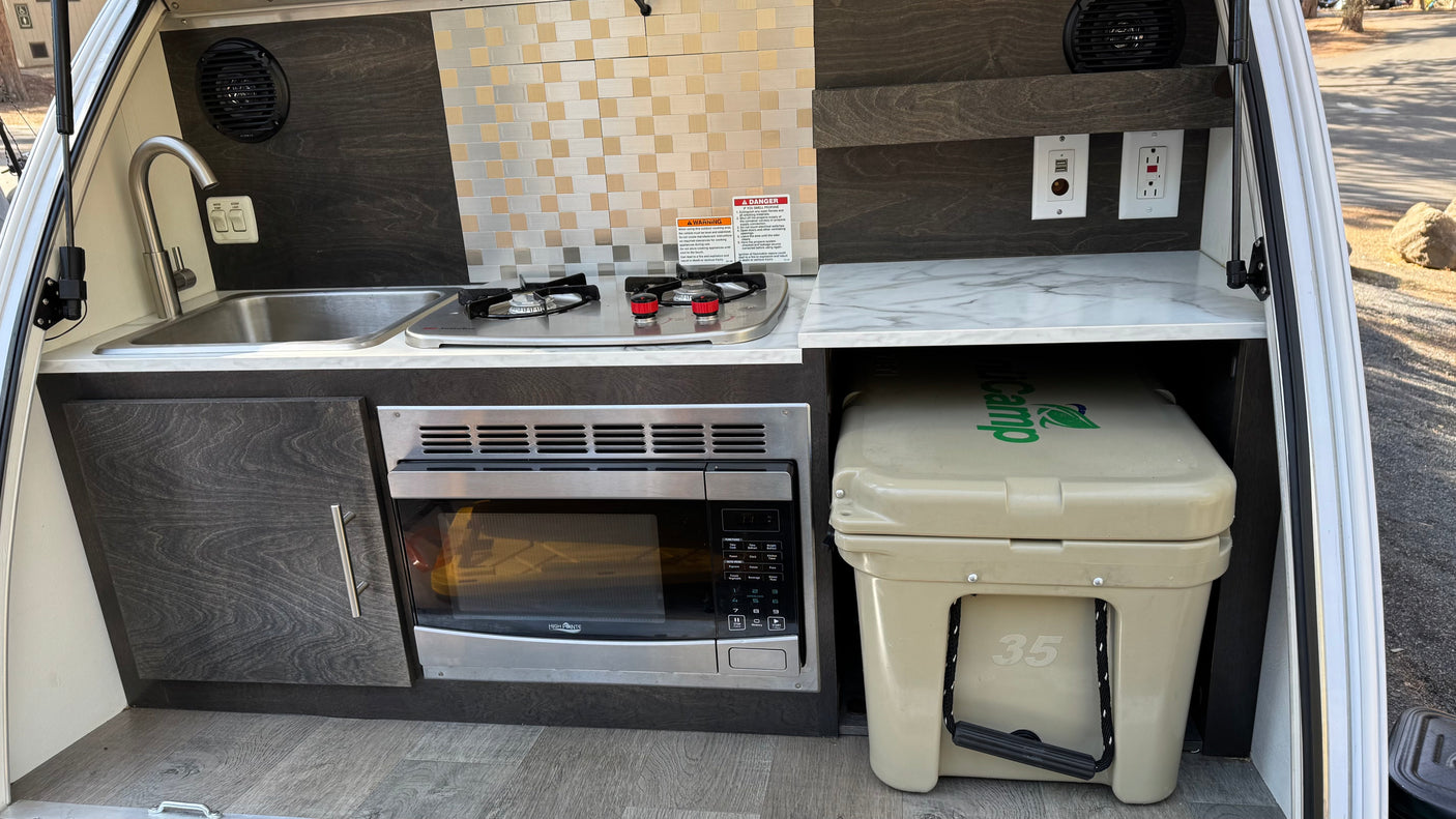 Compact RV rental outdoor kitchen with a sink, stove, microwave, and cooler, perfect for camper adventures.
