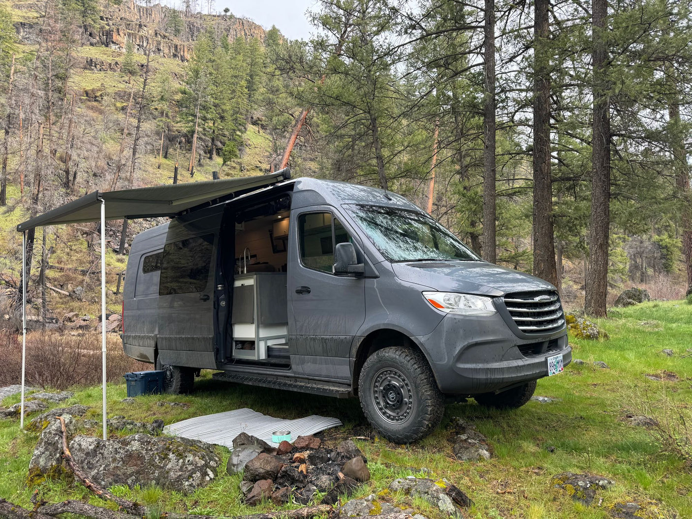 Spacious gray RV rental van with awning extended at a lush, rocky forest campsite, ideal for nature trips.