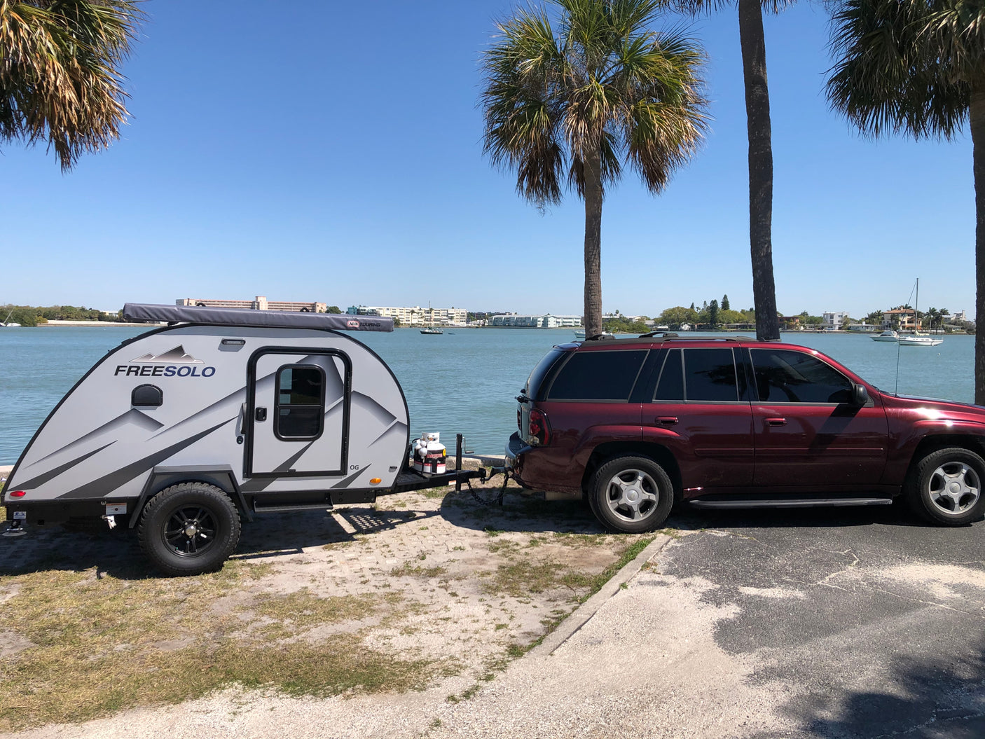 A grey Freesolo teardrop RV rental hitched to a red SUV, parked by a sunny waterfront with palm trees and boats in the distance.
