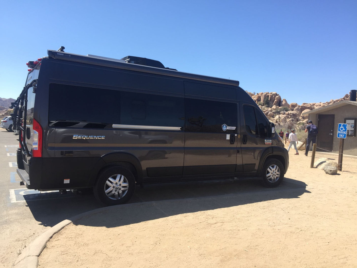 Dark gray RV rental camper van with awning & bike rack parked in a sunny, rocky desert landscape near restrooms.