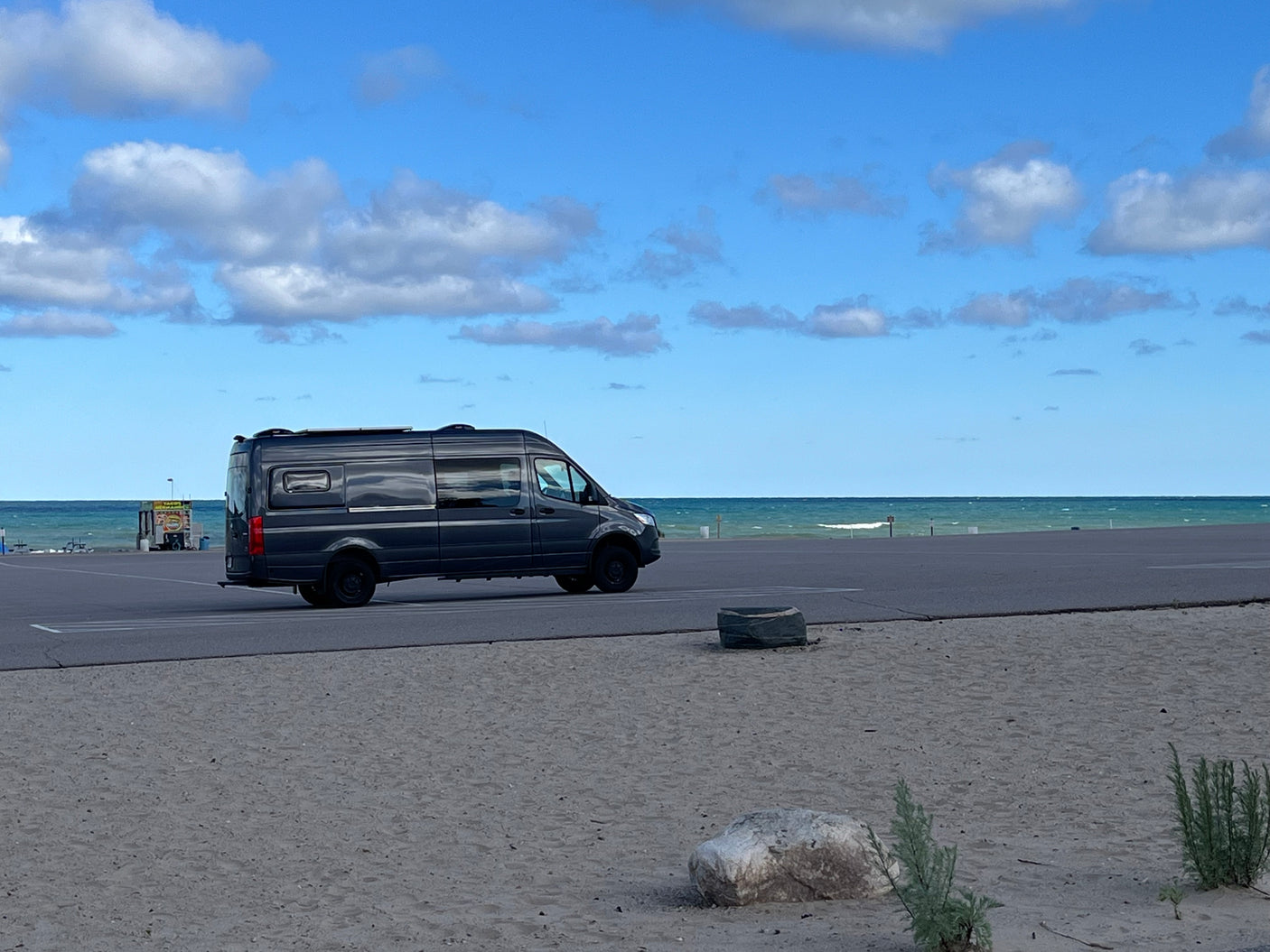 A dark grey RV rental is parked on an asphalt lot by a sandy beach with blue water and clouds.
