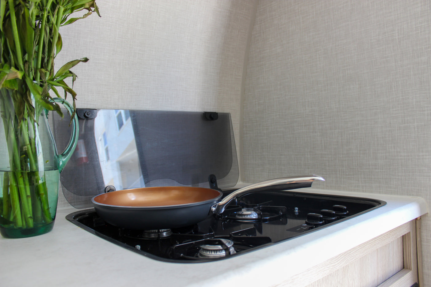 An RV rental kitchen features a compact stove with a frying pan, and a glass pitcher of green stalks on the white countertop.