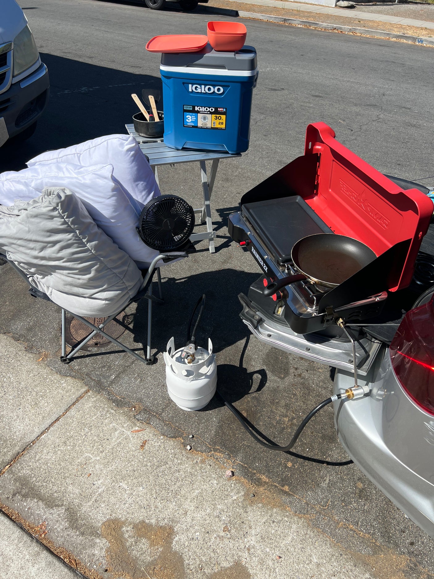 RV rental outdoor cooking station: a red portable stove, propane tank, blue Igloo cooler, fan, and bedding on asphalt.