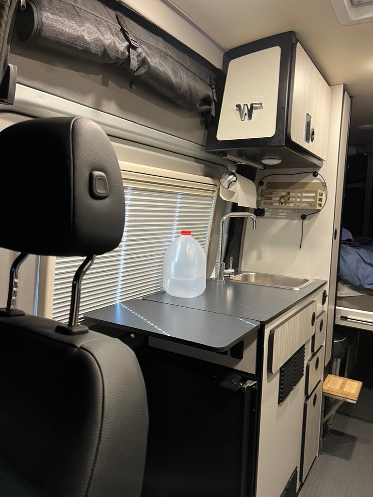 This RV rental features a compact kitchen with a sink, counter, and fridge. Overhead storage & window blinds visible. Ideal for travel.