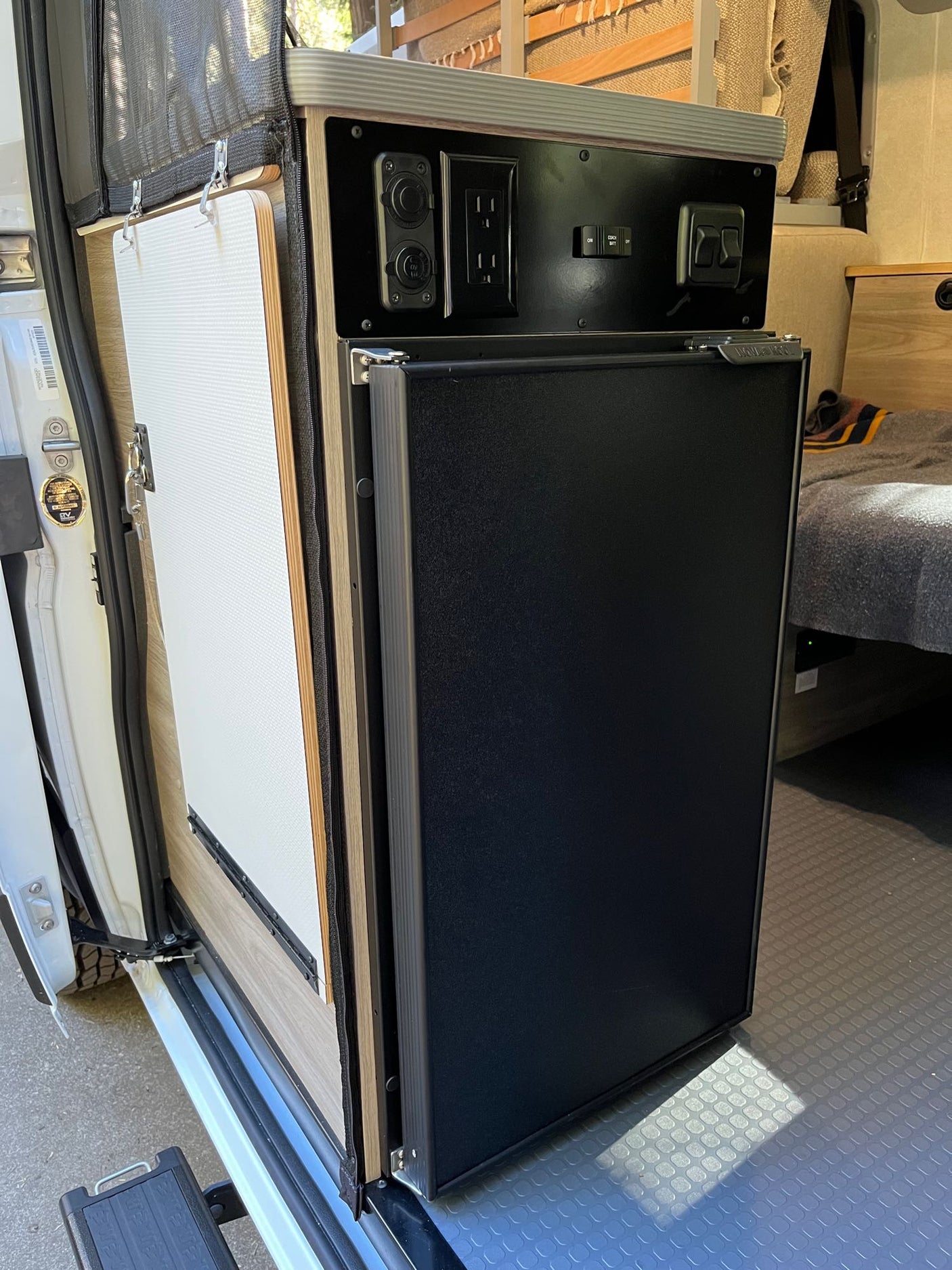 RV rental: Black refrigerator, control panel with outlets, and a pull-out table/door panel in a camper van interior.
