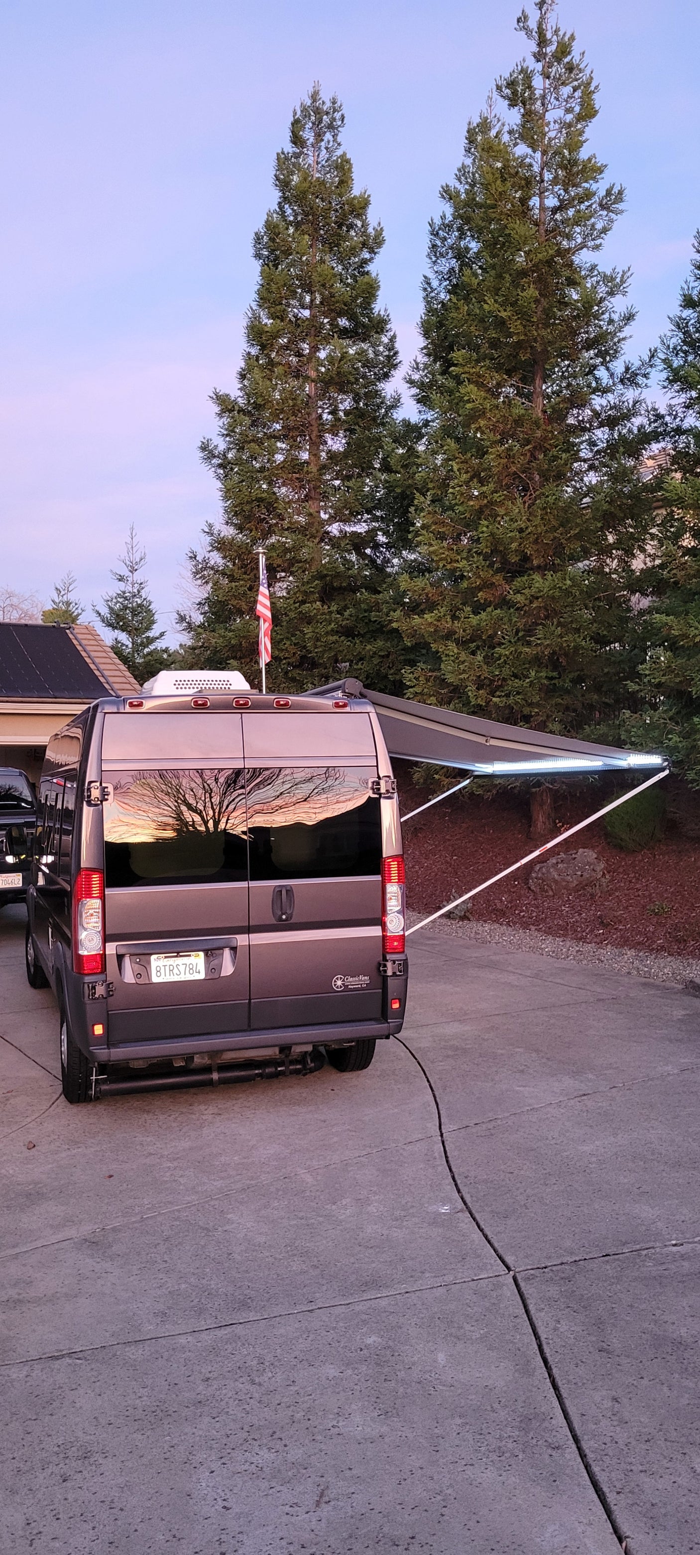 RV rental van with illuminated awning and flag. Sunset reflects in windows; parked on driveway. Ready for adventure.