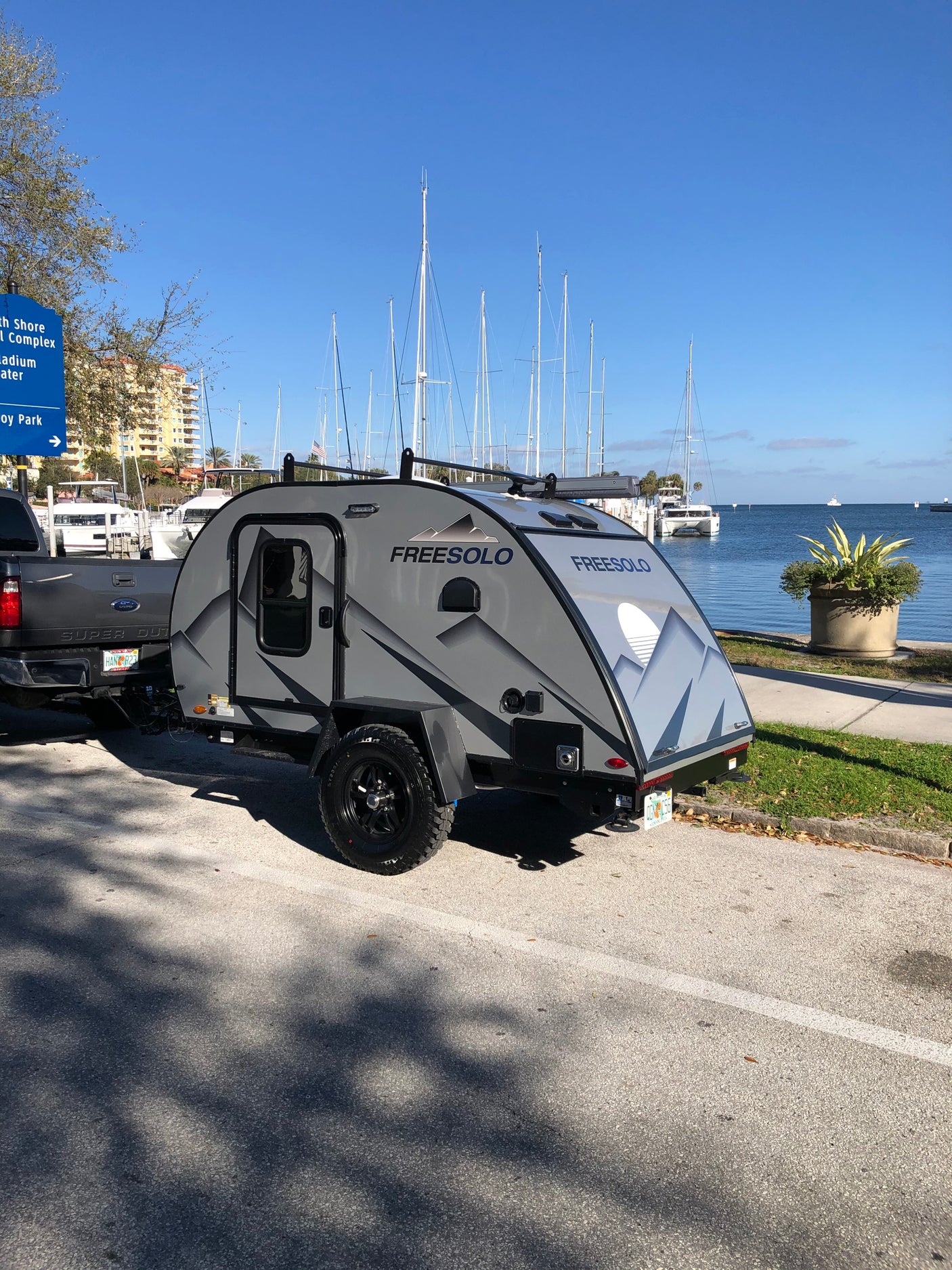 Grey Freesolo teardrop rv rental hitched to a truck, parked by a marina filled with sailboats. Ideal for outdoor adventures.