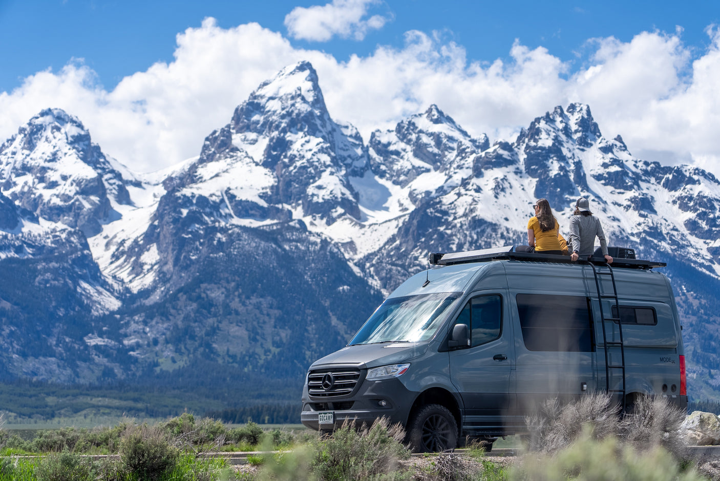 Couple on an RV rental enjoys majestic snow-capped mountains from their camper van roof.