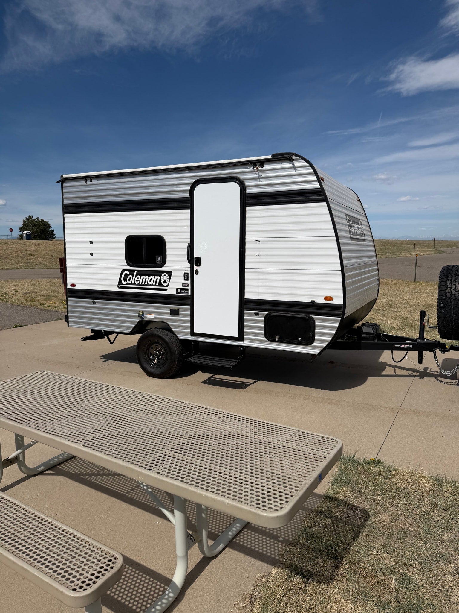Enjoying the outdoors with this Coleman rv rental. A small travel trailer is parked by a metal picnic table under a blue sky.