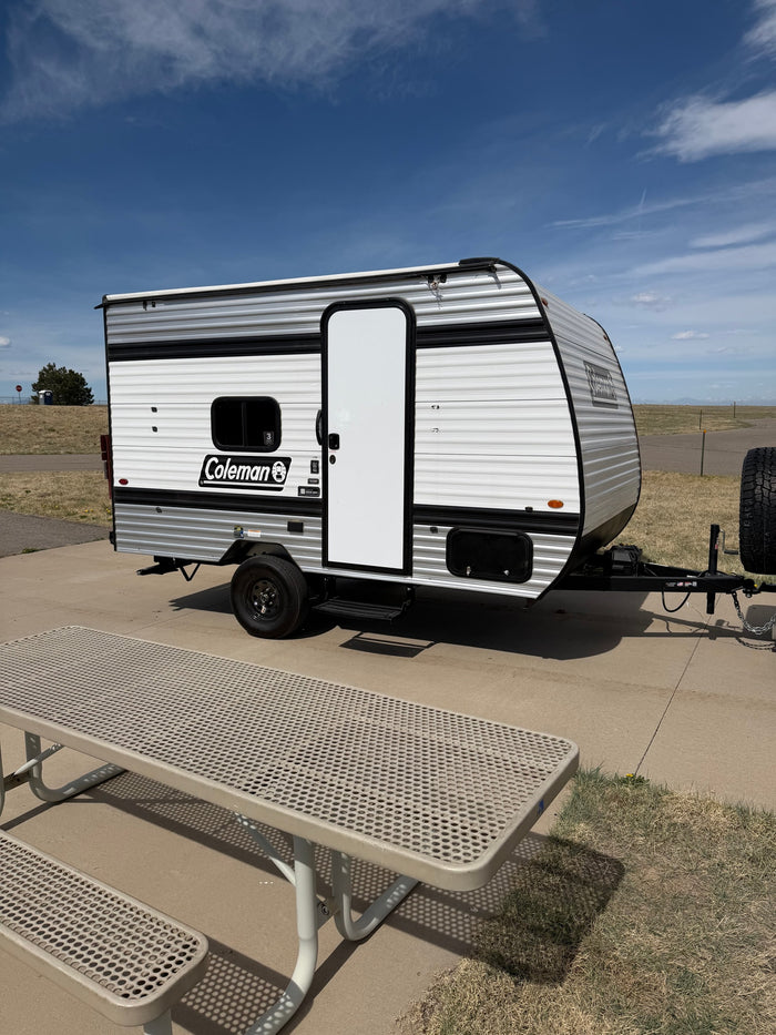 Enjoying the outdoors with this Coleman rv rental. A small travel trailer is parked by a metal picnic table under a blue sky.