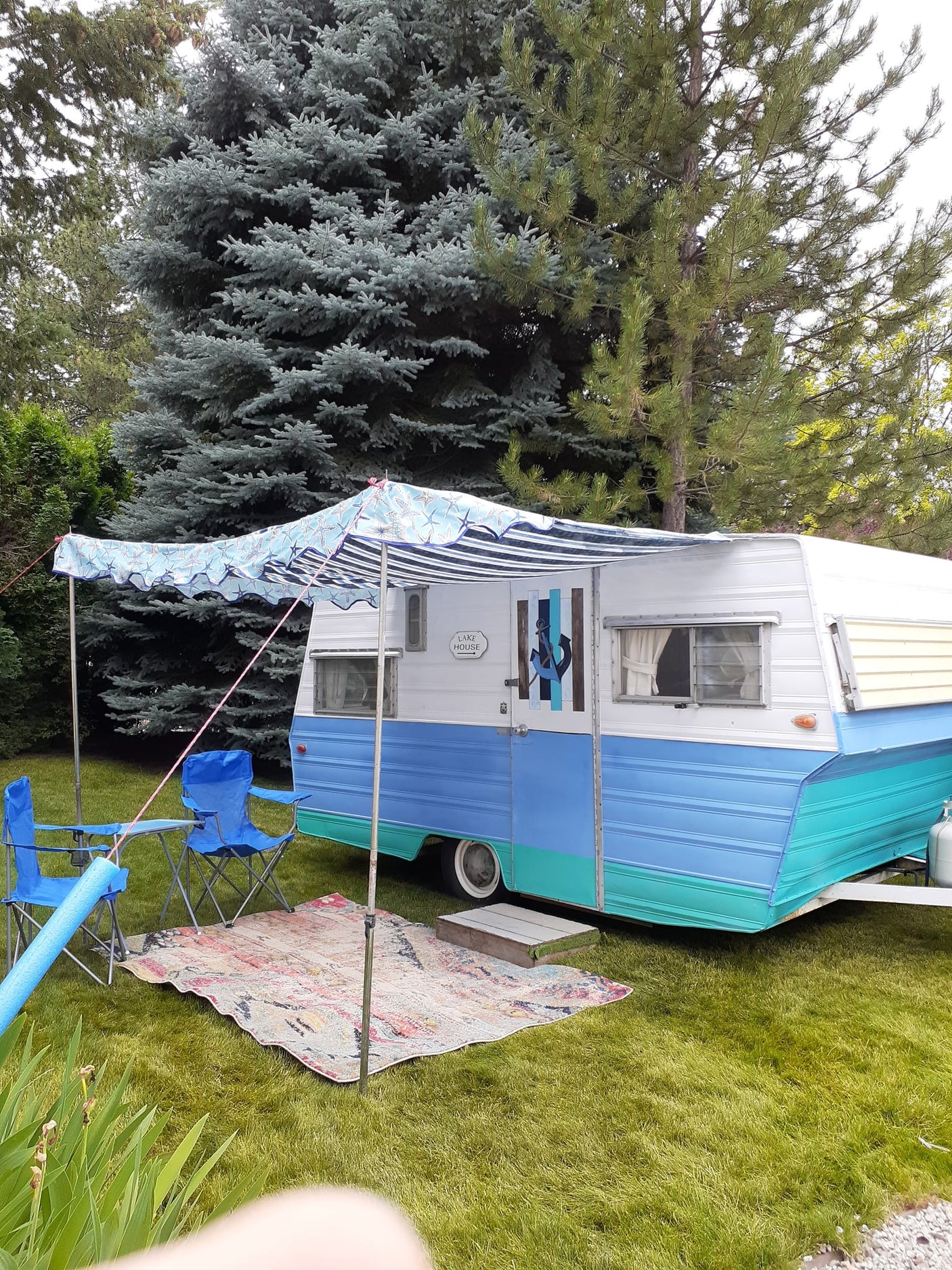 This colorful teal & blue RV rental is set up for lake house camping, with an awning, chairs, and rug on the grass.