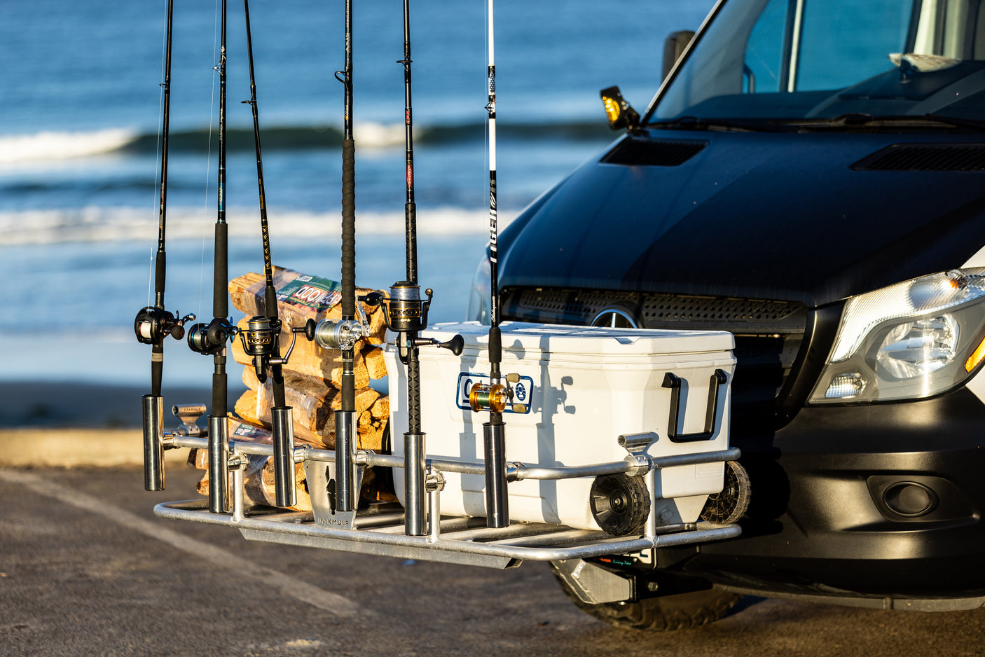 An RV rental van equipped for a beach fishing trip, with rods, cooler, and firewood on its front carrier.