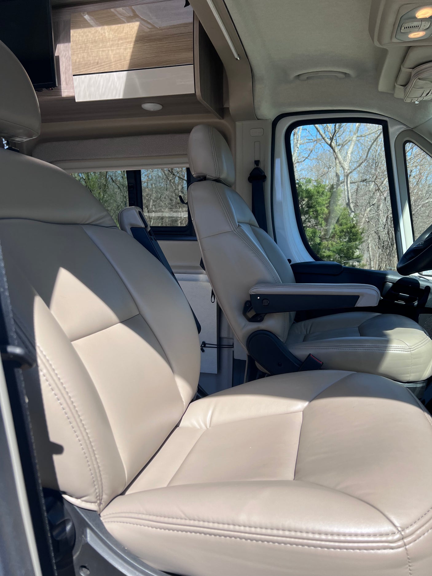 Front cabin of an RV rental with comfortable, light-colored captain's chairs, overhead storage, and a bright outdoor view.