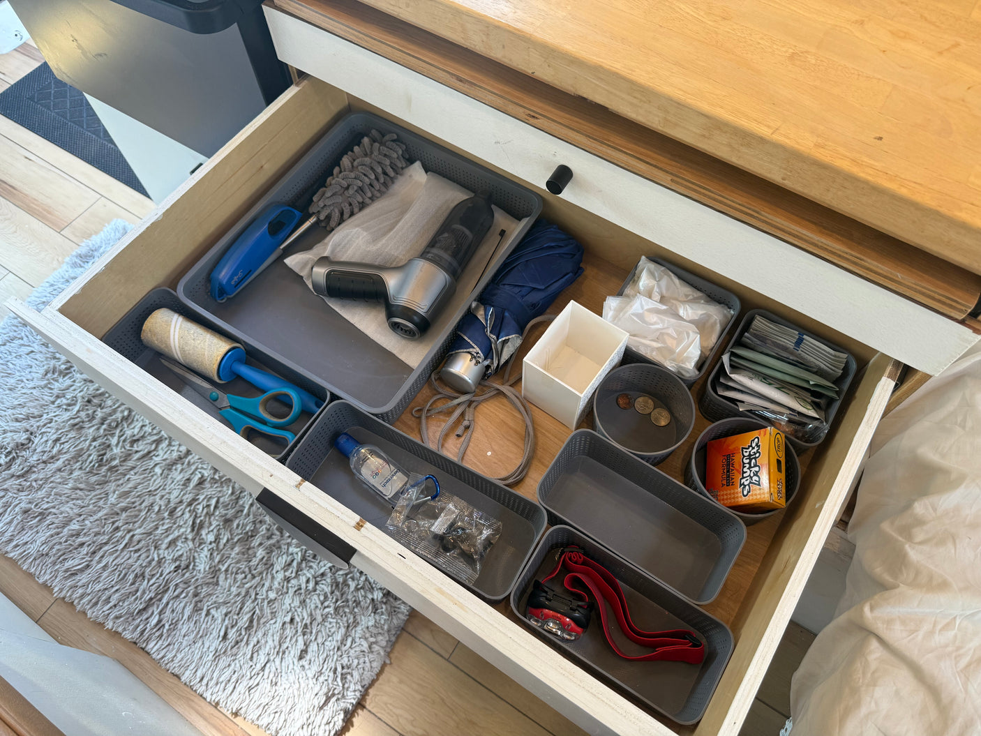 Inside an RV rental drawer: travel supplies like a mini vacuum, lint roller, umbrella, and Sticky Bumps wax neatly stored in gray bins.