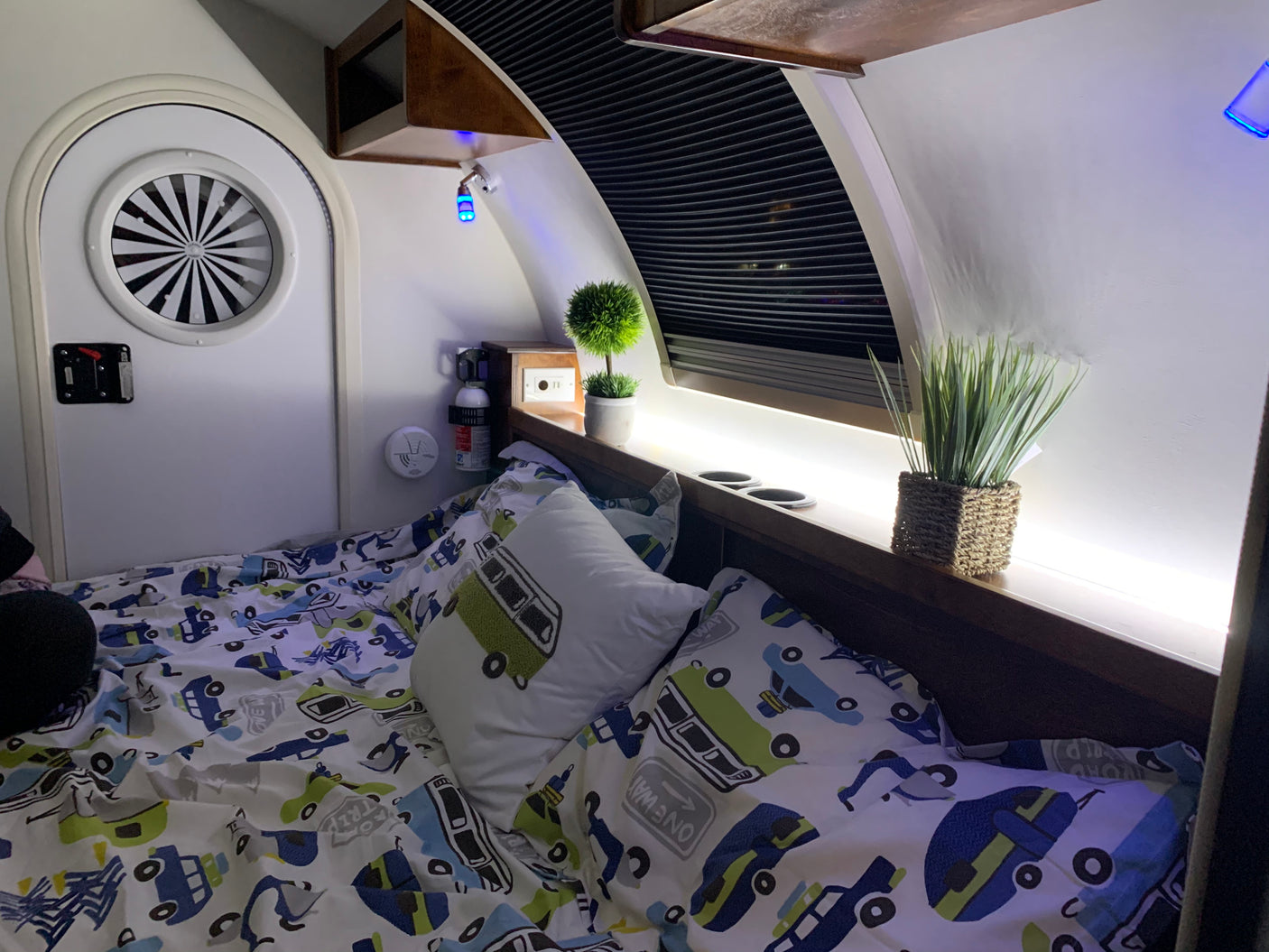 Cozy RV rental sleeping area: bed with car-themed bedding, circular window, blue accent & LED lighting, safety gear.