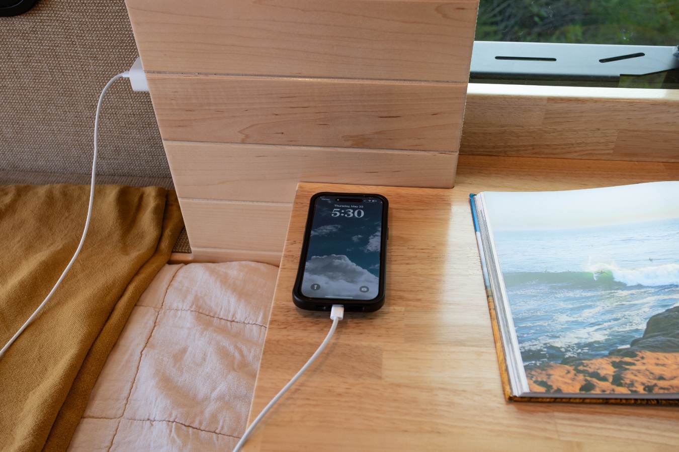RV rental interior: Phone charging on a light wood surface next to an open book showing ocean waves, ideal for travel.