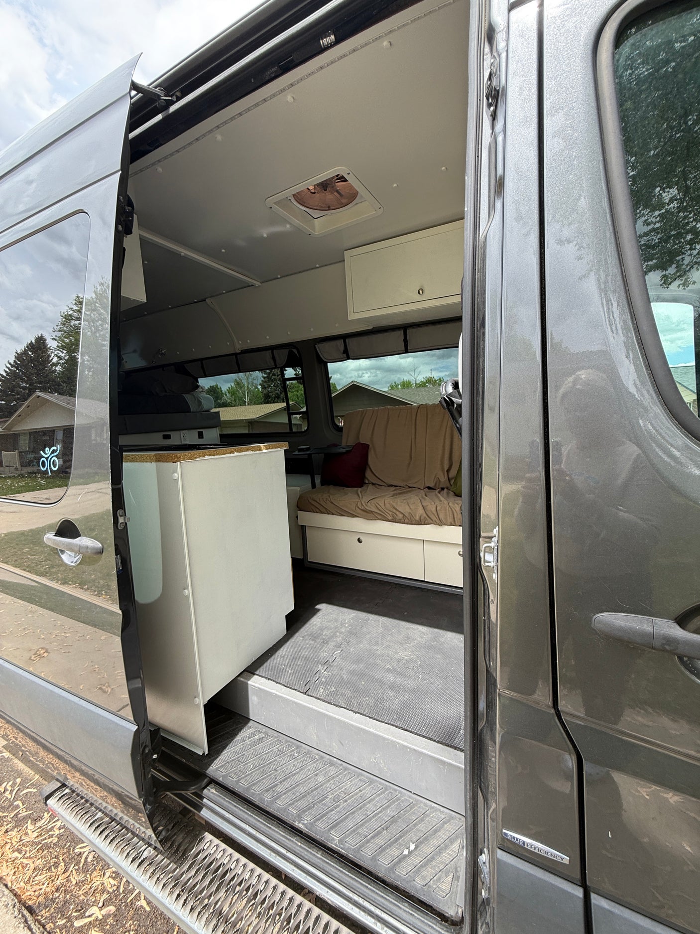 RV rental: Interior view of a grey converted camper van through its open side door, revealing a bed, storage, and ceiling fan.