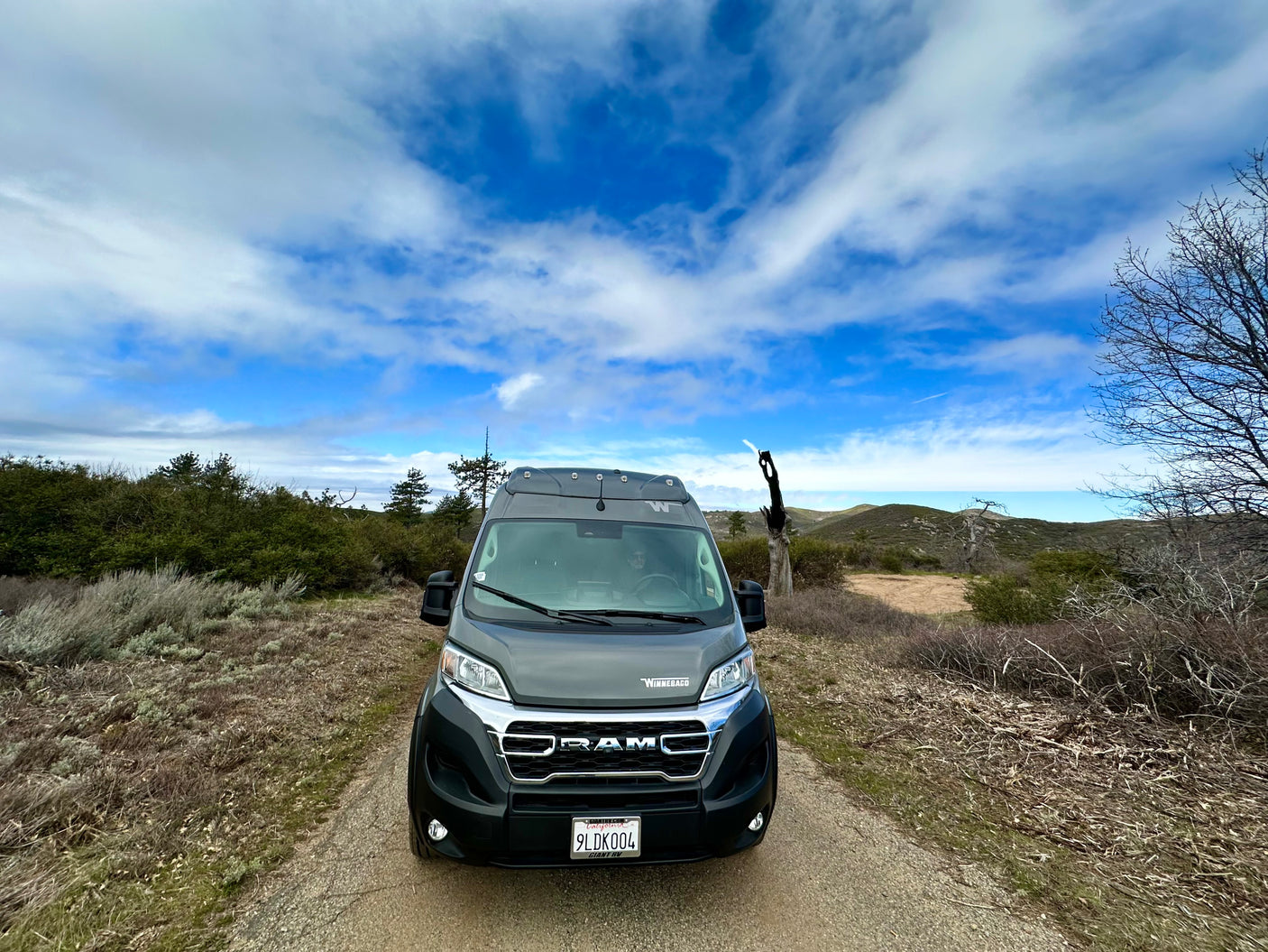 Front view of a gray Winnebago campervan RV rental on a dirt path, surrounded by hills and cloudy blue sky.