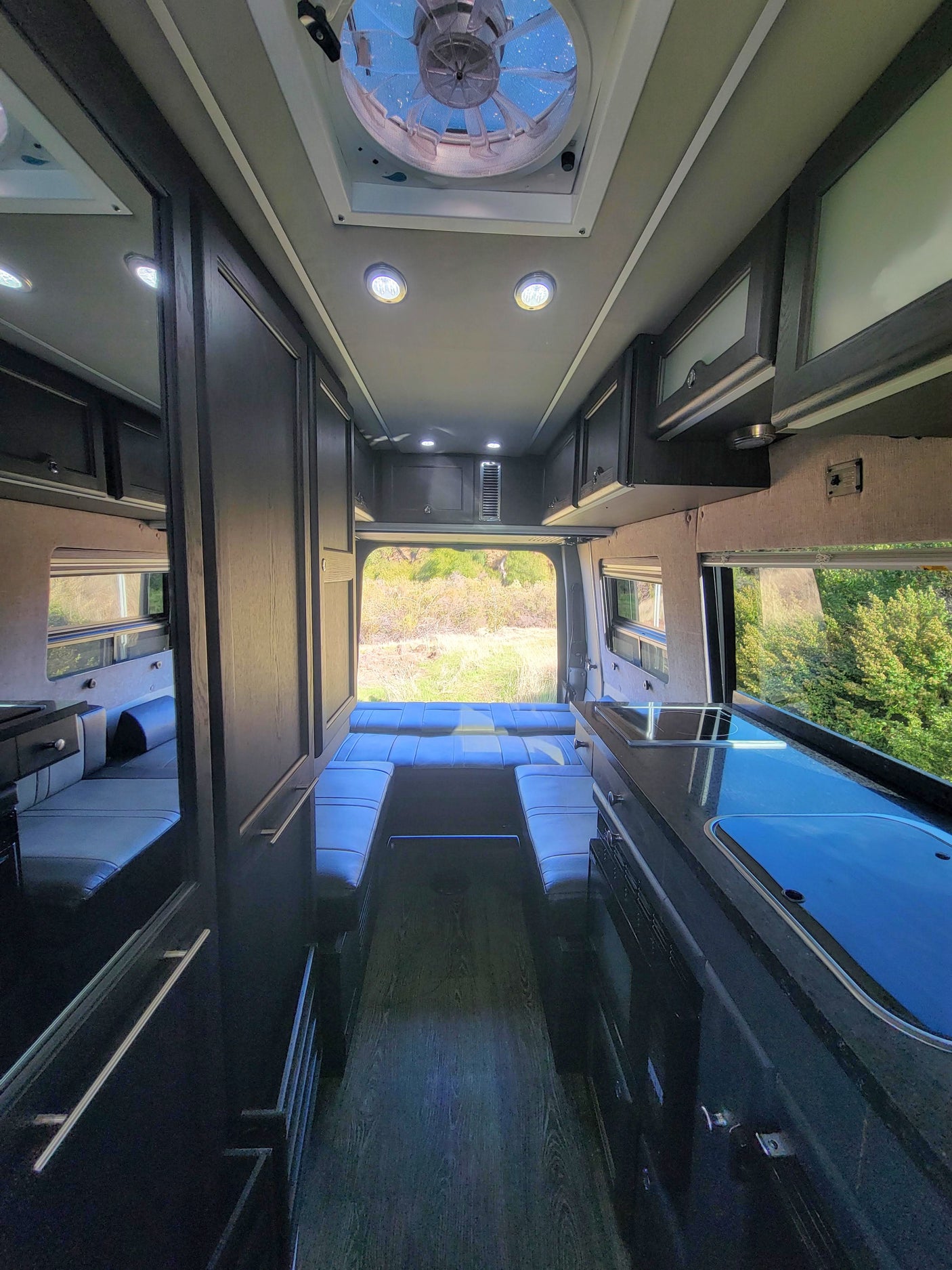 Spacious RV rental interior with kitchenette, lounge/bed, and overhead storage, looking out to nature.