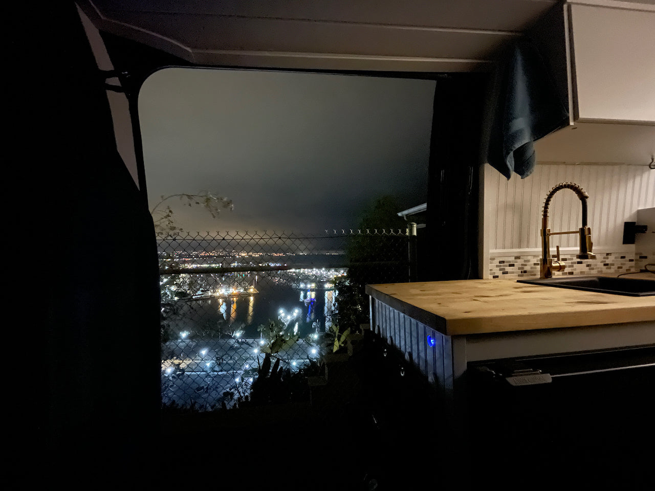 Night view from an RV rental: a kitchen with gold faucet overlooks city lights, a fenceline, and water. Mobile living awaits!