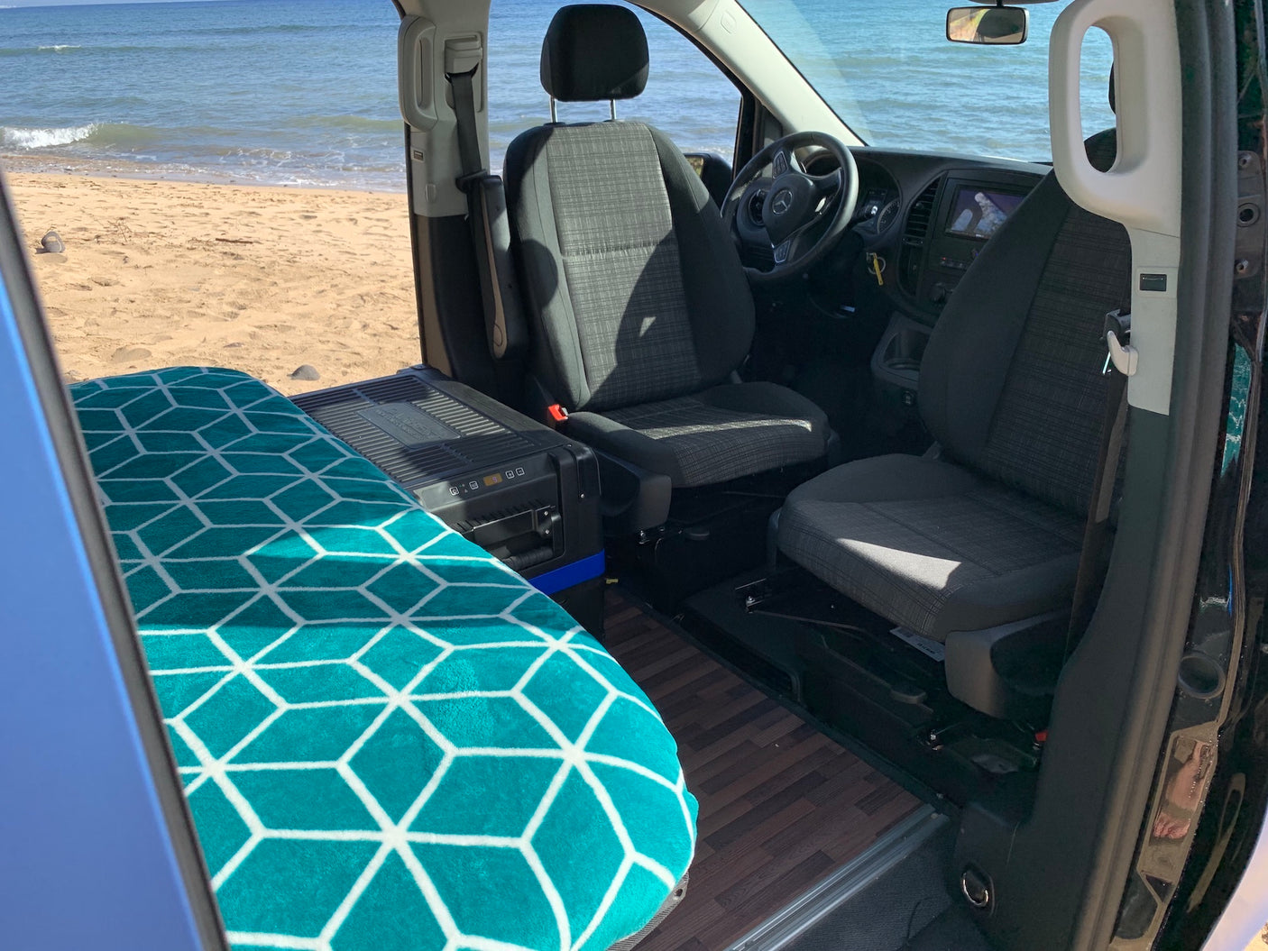RV rental interior shows a cozy bed, portable fridge, and front seats, all set against a stunning beach and ocean view.