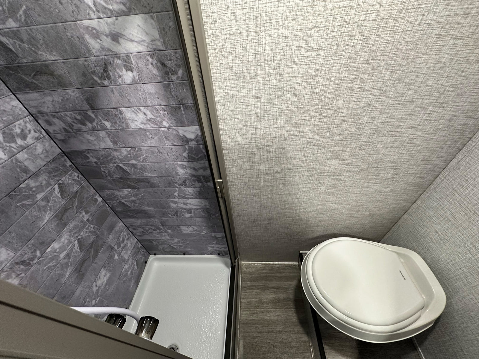 Compact RV rental bathroom: view of a grey marble-tiled shower with a white pan, and a white toilet.