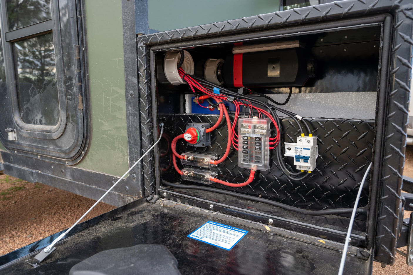 An rv rental's open diamond plate compartment reveals its electrical power system: battery, fuse panel, and wiring.
