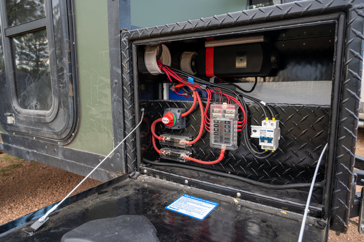 An rv rental's open diamond plate compartment reveals its electrical power system: battery, fuse panel, and wiring.