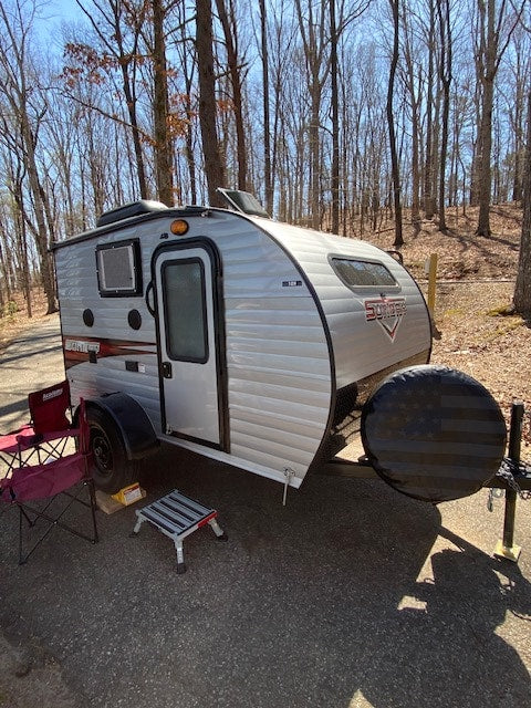 Silver teardrop rv rental travel trailer parked in a sunny wooded campground. Features a spare tire with an American flag cover.|Scenic lake view with a white pickup towing an RV rental. Perfect for outdoor adventures!|This compact RV rental