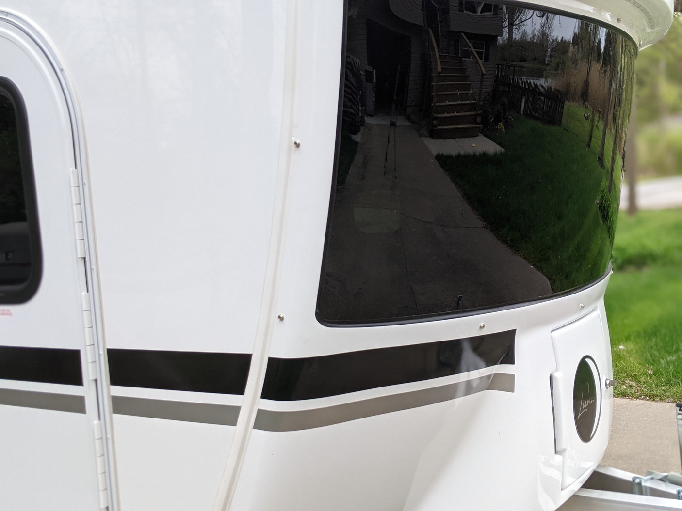 White travel trailer RV rental: its side features a large dark window reflecting a home/grass, black stripes, and a 'Luna' emblem.