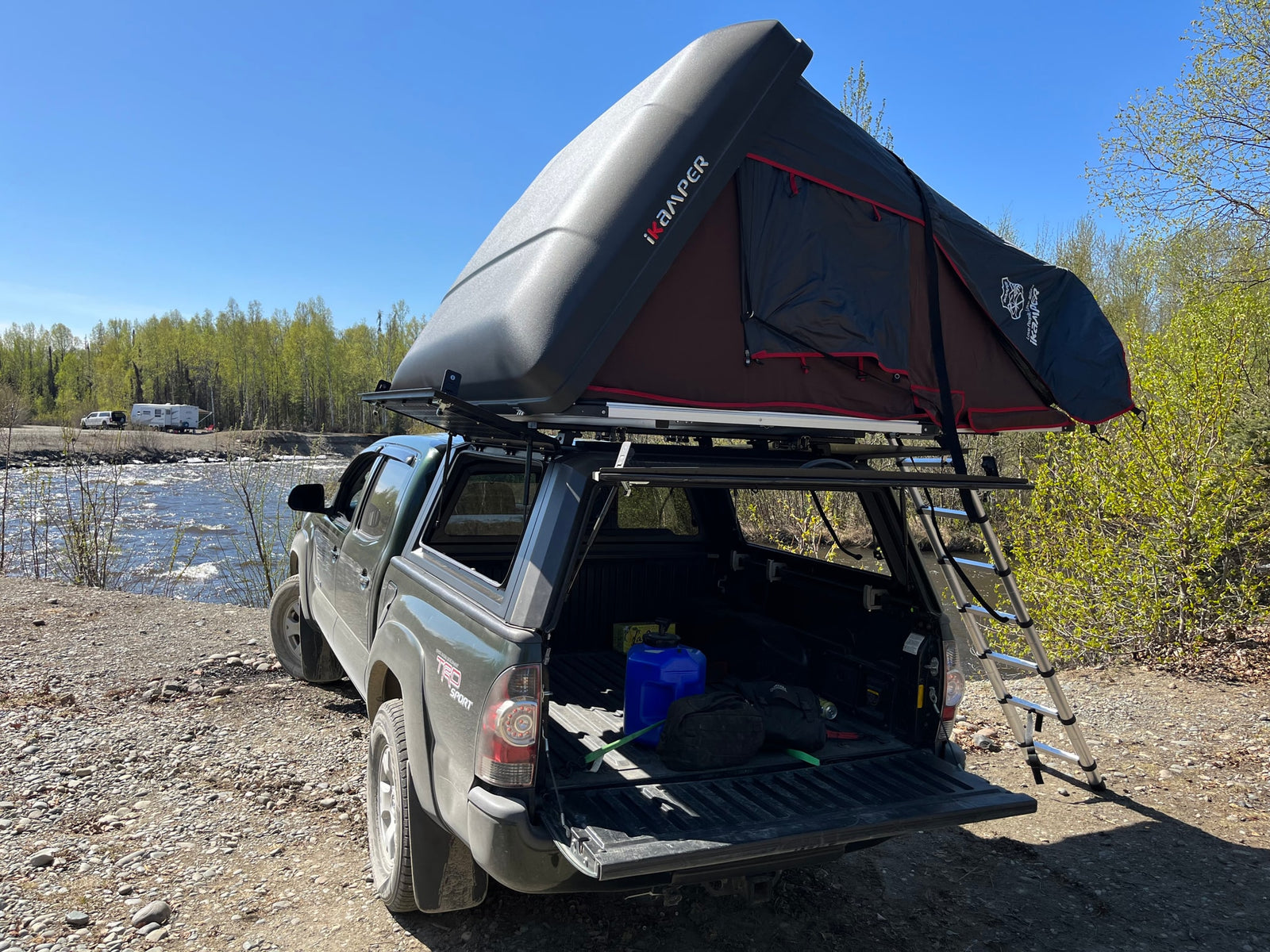 A truck with an iKamper rooftop tent parked by a river, with an RV in the distance. Perfect for outdoor camping adventures or an RV rental alternative.