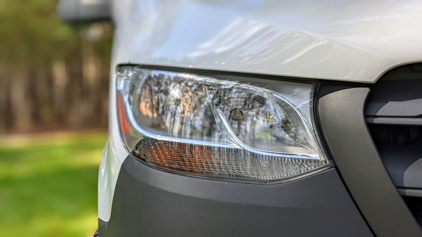 White RV rental van: close-up of its front right side, showing the headlight and black bumper. Outdoors in daylight.