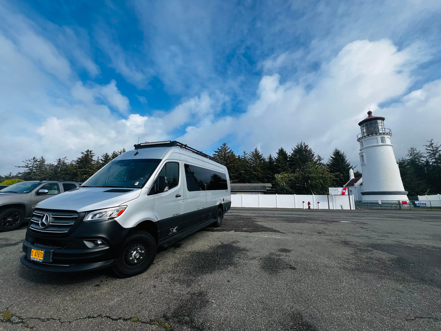 Silver Mercedes-Benz Sprinter RV rental parked at a historic lighthouse, nestled among evergreen trees under a cloudy blue sky.