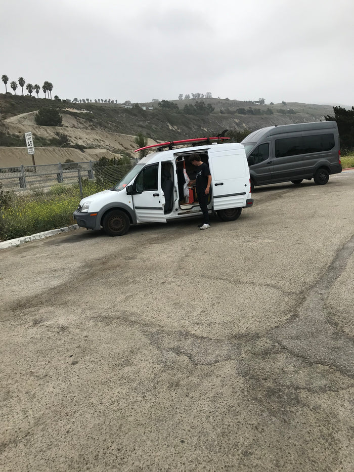 Person organizing inside a white van with a surfboard, alongside a gray van on a scenic hillside. Ideal for an **RV rental** trip.