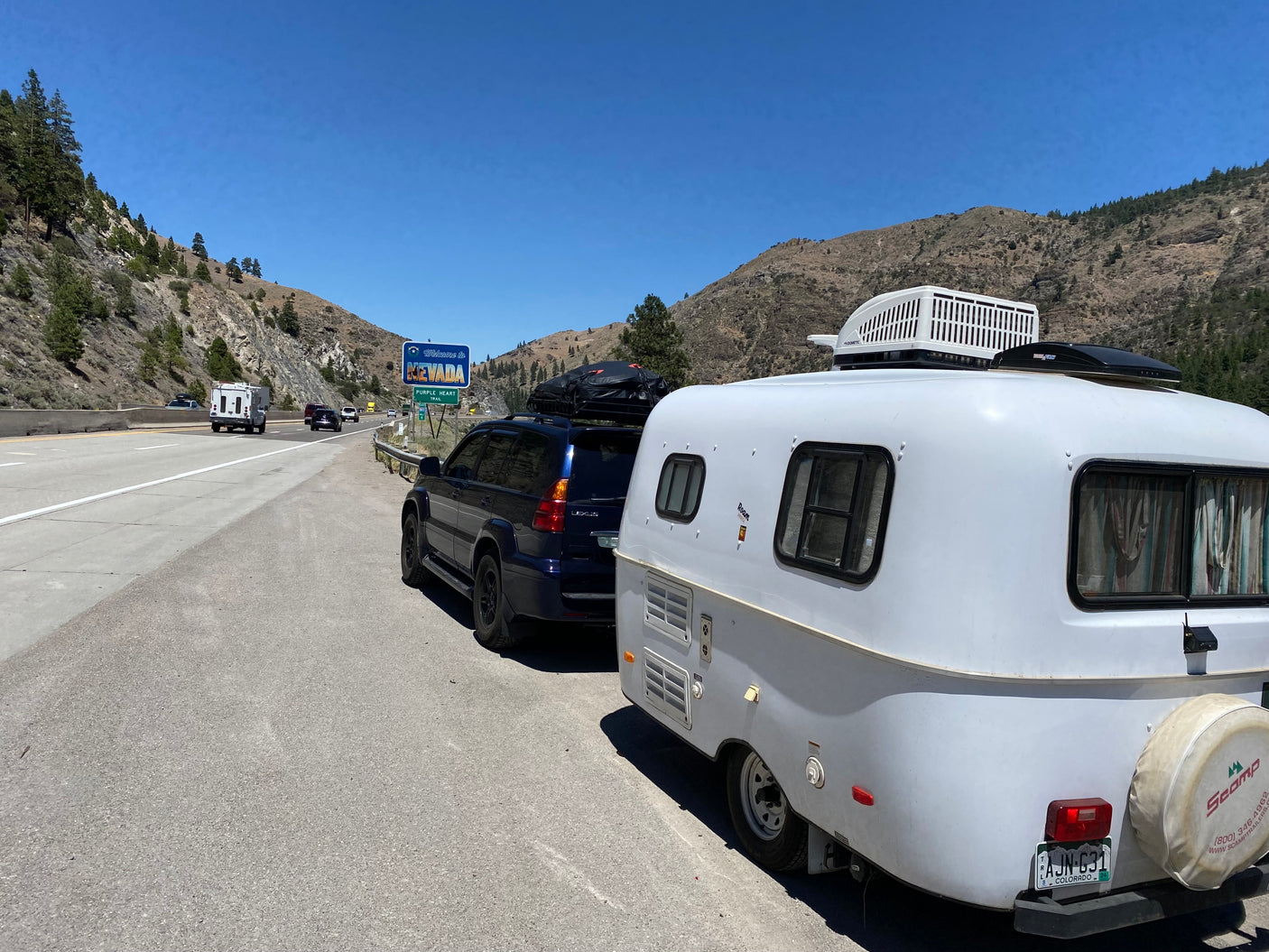 White Scamp rv rental and Lexus SUV parked on a Nevada highway next to the state sign, ready for mountain adventures.