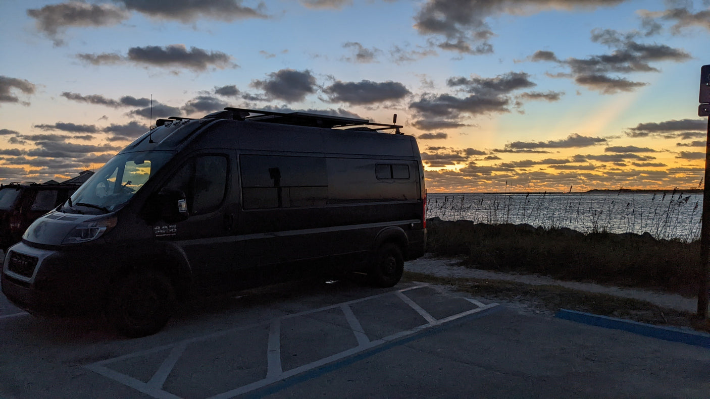 A dark RV rental campervan is parked seaside at sunset, showcasing a beautiful sky. Perfect for travel exploration.