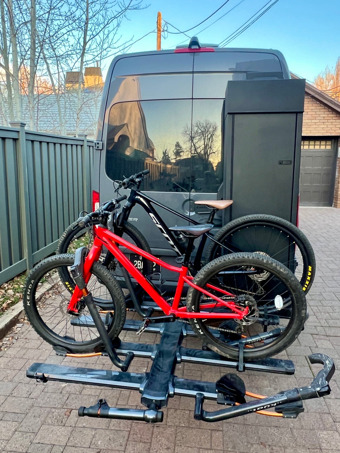 Dark gray RV rental with two mountain bikes (red & black) on a rear hitch rack, plus a storage box, parked on pavers.