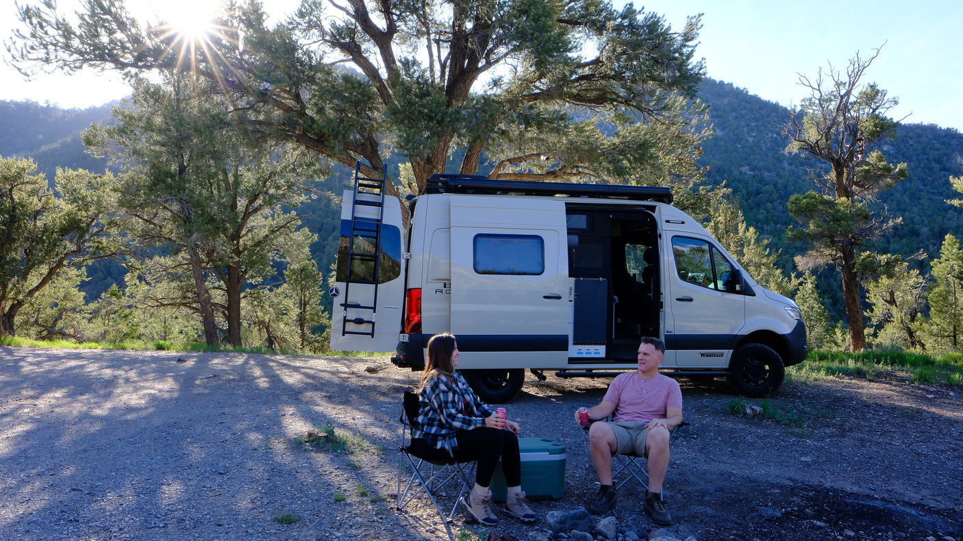 Enjoying nature in an RV rental! A couple relaxes in camping chairs next to their camper van amidst trees and mountains.