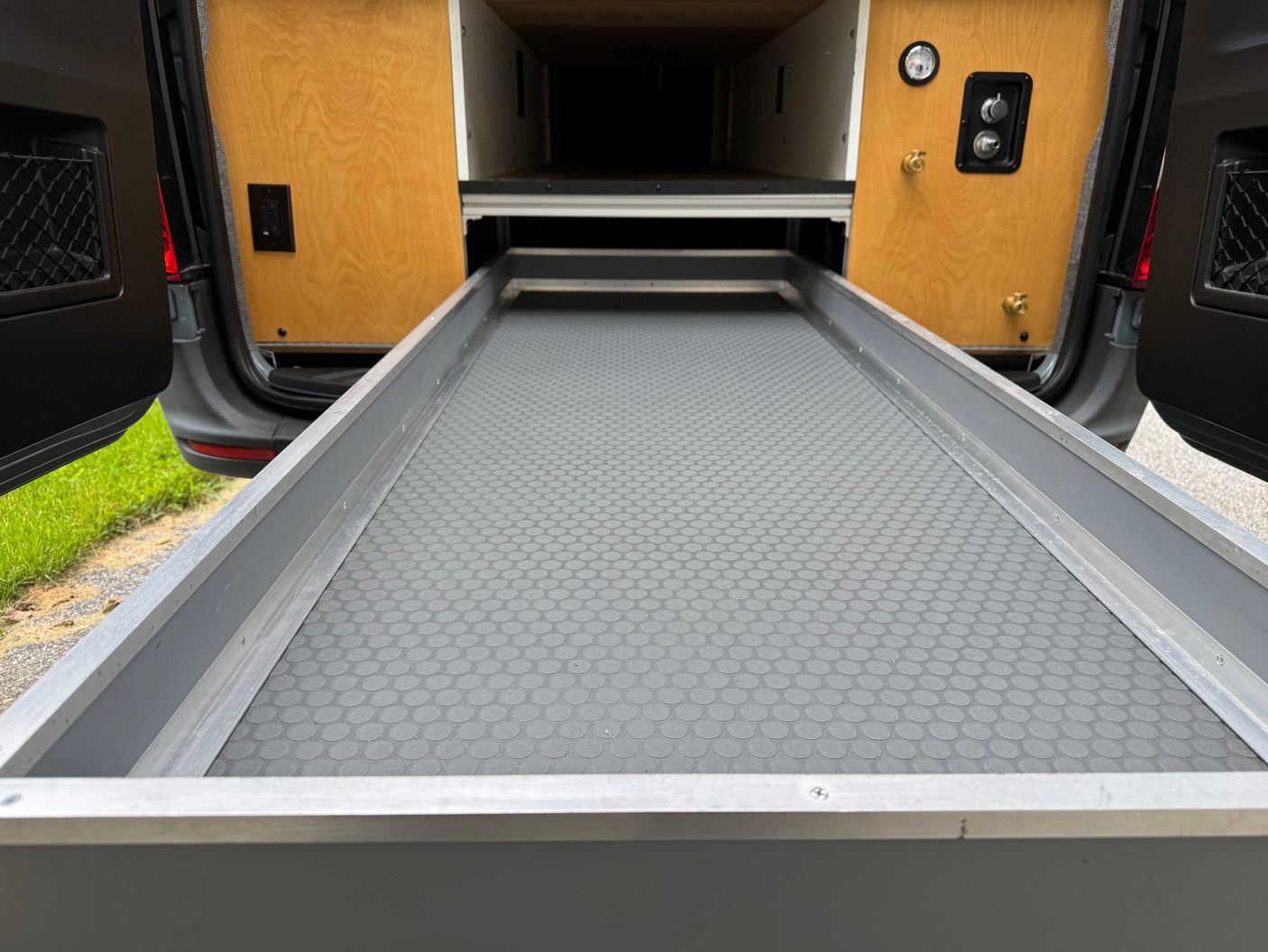 RV rental: Camper van's extended pull-out cargo tray with a textured mat, flanked by wood panels containing a gauge and water connections.