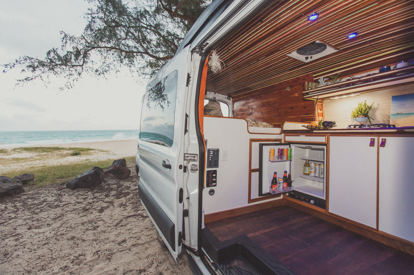 Beachside living: Explore this custom RV rental, a camper van with open door revealing fridge, kitchen, and bed, parked by the ocean.