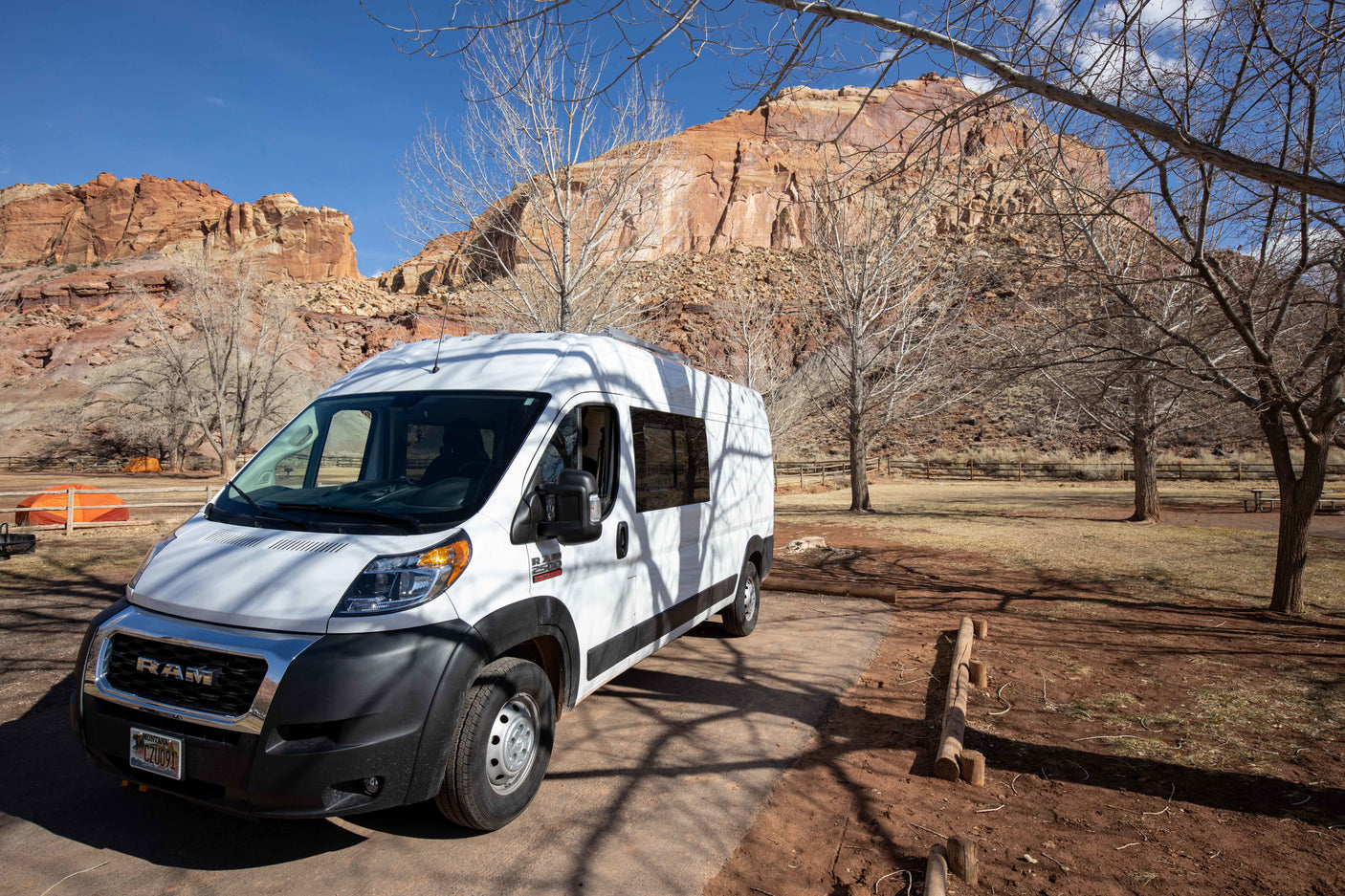 White RV rental camper van parked in a scenic desert campsite with striking red rock formations and bare trees.