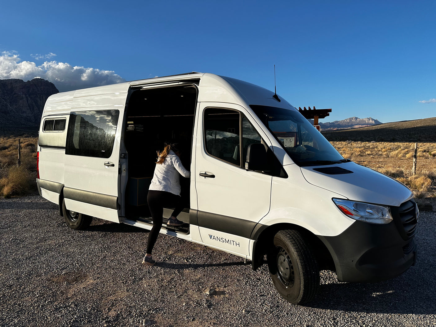 Ready for adventure! A person steps into a white VanSmith RV rental van, parked in a scenic desert landscape.