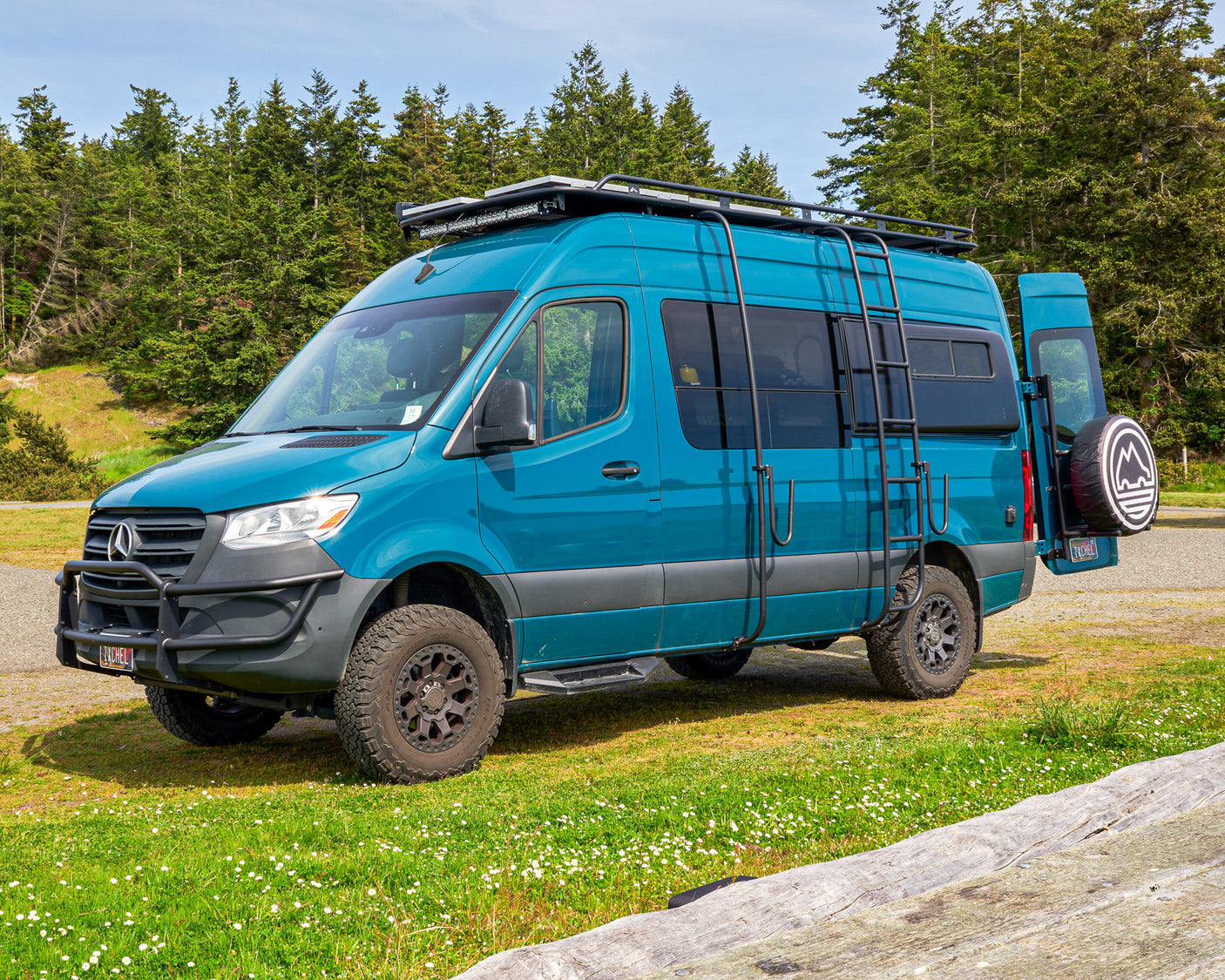 This teal, rugged Mercedes Sprinter adventure van, equipped for off-road travel, is a top choice for an RV rental, parked by a forest.