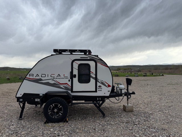 Radical Bushwhacker travel trailer RV rental, equipped for adventure, sits on gravel under a cloudy sky, mountains in distance.