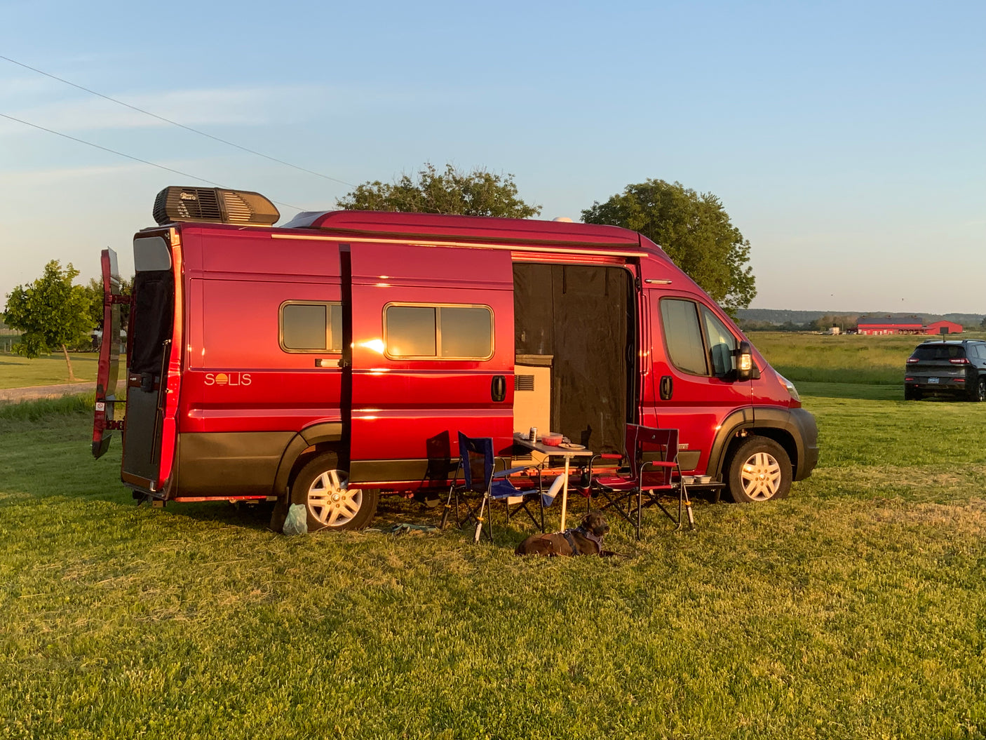 This red Winnebago Solis RV rental offers a perfect camping setup. Parked in a field at sunset, with chairs, table, and a dog.