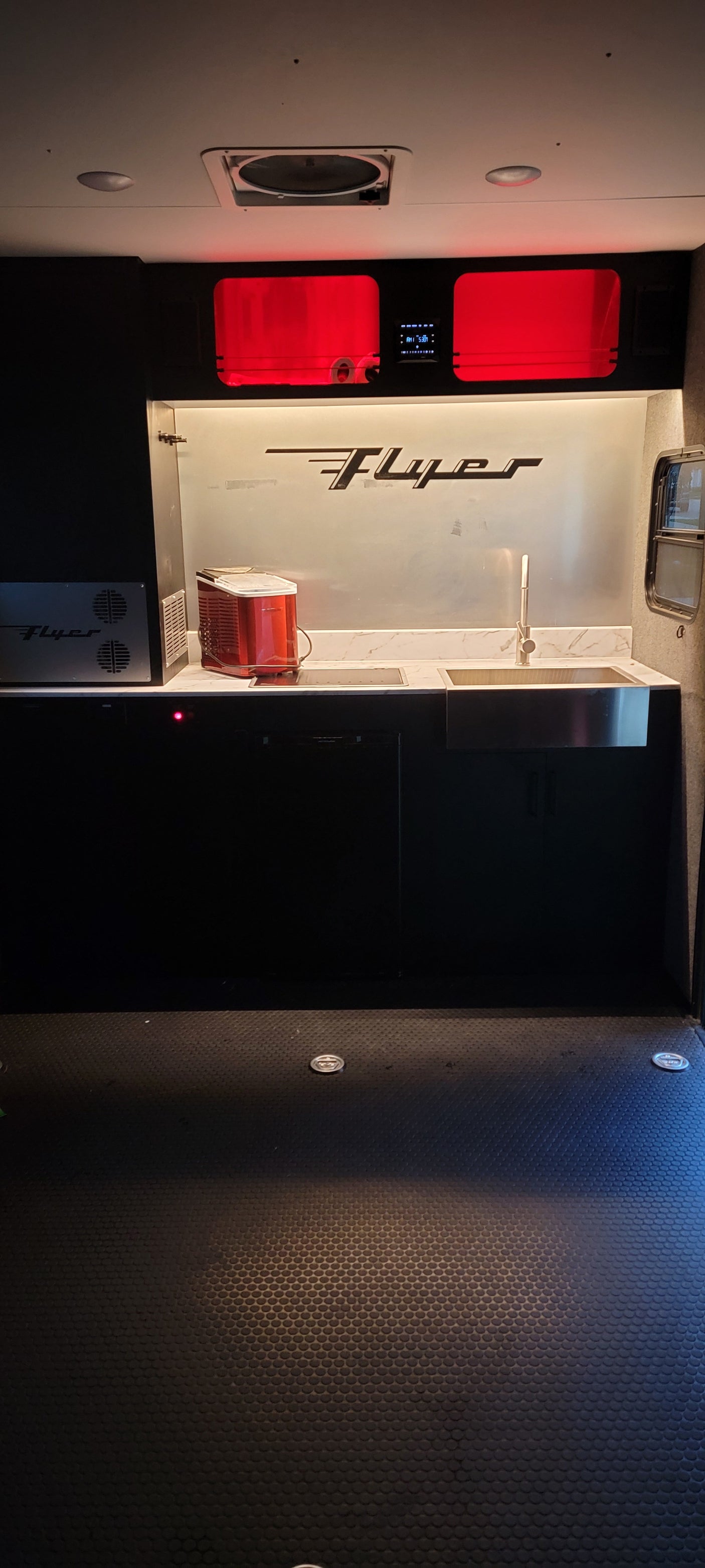 Compact RV rental interior featuring a modern kitchenette with sink, cooktop, red ice maker, and overhead red lighting.