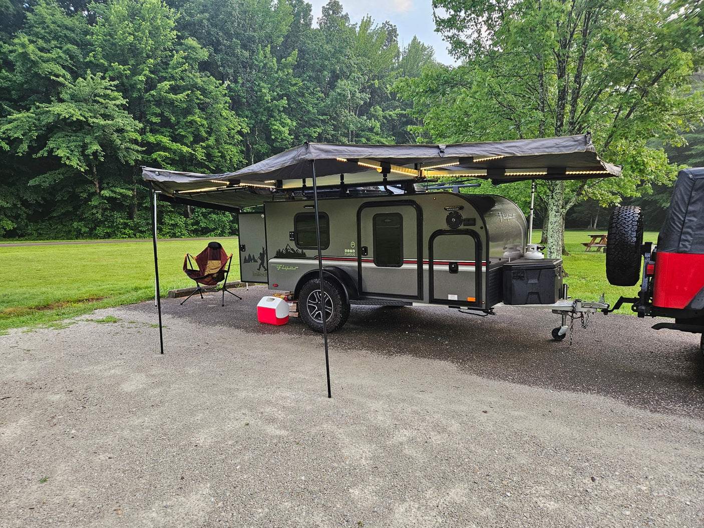 An RV rental teardrop camper with illuminated awning, chair, and cooler at a serene green campsite. Perfect for outdoor adventures!
