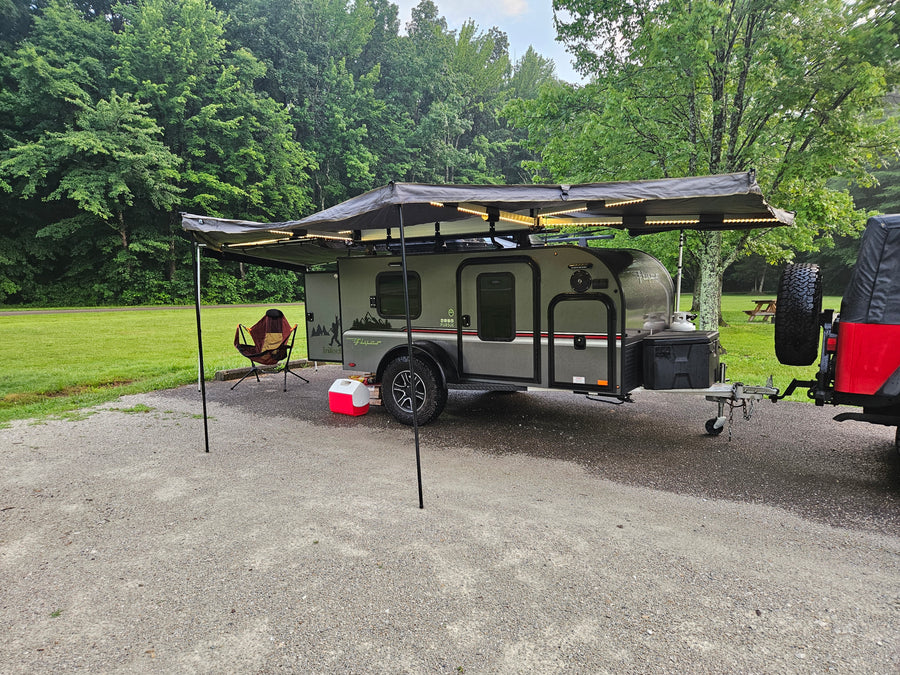 An RV rental teardrop camper with illuminated awning, chair, and cooler at a serene green campsite. Perfect for outdoor adventures!